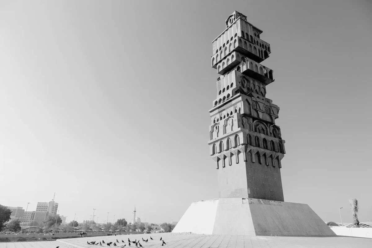 A Jeddah landmark tower rising above an open plaza, with pigeons in the foreground and the city behind